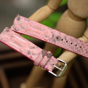 Könnte beinhalten: Rosa Uhrenarmband mit Schlangenmuster. Das Armband hat eine silberne Schnalle und mehrere Löcher zur Anpassung. Das Leder hat ein rosa und braunes Farbschema. Das Uhrenarmband wird vor einem verschwommenen Hintergrund gezeigt.