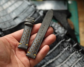 Könnte beinhalten: Zwei graue Lederarmbänder mit gelber Naht und einem perforierten Design. Die Armbänder werden in einer Hand gehalten.