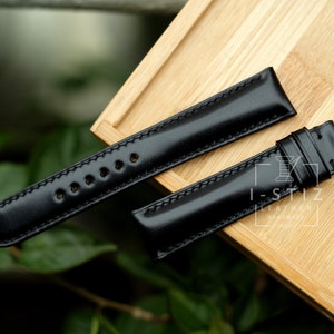 May include: Two black leather watch straps with stitching detail. The straps are laid out on a wooden surface.