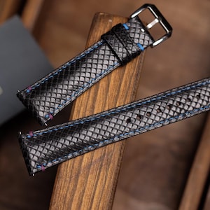 Peut inclure: Bracelets de montre noirs avec un motif texturé et des coutures bleues. Les bracelets sont en cuir et ont une boucle argentée. Les bracelets de montre sont présentés sur une surface en bois.