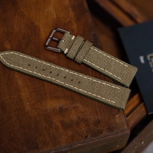 May include: An olive green canvas watch strap featuring white contrast stitching along the edges. The strap includes a silver-toned metal buckle and two matching canvas keepers. The material has a textured, woven appearance with visible stitching.