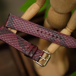 May include: Two-piece watch strap in a burgundy and black snakeskin pattern. The leather strap has a silver-colored buckle and is displayed on a wooden mannequin. The watch band is a stylish accessory.