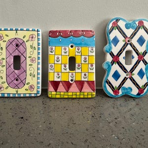Peut inclure: Trois plaques d'interrupteur en céramique rectangulaires avec des motifs peints à la main. La plaque de gauche a un fond jaune avec des roses roses et un centre violet. La plaque centrale présente une grille jaune avec de petites fleurs. La plaque de droite a un motif en losange bleu avec des roses rouges.