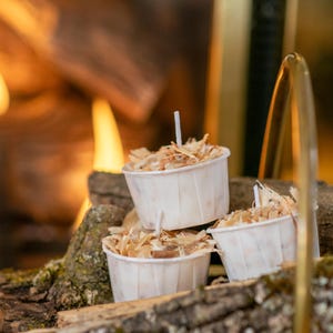 May include: Three small, white paper cups filled with wood shavings and a small white wick, stacked on a piece of wood. The background shows a fireplace with flames and a gold-coloured metal object.