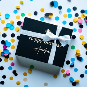 May include: A black gift box with a white ribbon and the text "Happy Birthday Amelia" in gold. The box is surrounded by colorful confetti in shades of blue, yellow, pink, and black, creating a festive birthday theme.