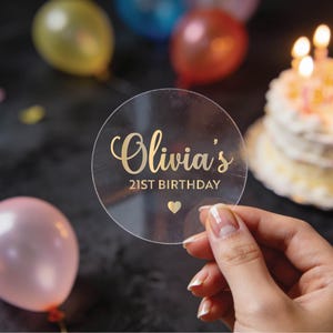 May include: A clear, round acrylic sign with gold lettering that reads "Olivia's 21st Birthday" and a small heart. The sign is held in front of a birthday cake with lit candles and colorful balloons.