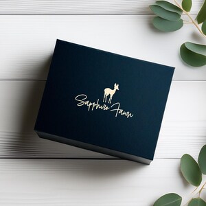 May include: A navy blue square box with a gold logo and text that reads "Sapphire Fawn". The box is on a white wooden surface with green leaves in the background. The logo features a deer.