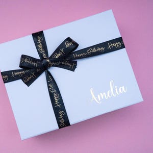 May include: A white gift box tied with a black ribbon printed with "Happy Birthday" in gold. The box is personalized with the name "Amelia" in gold script. The background is a pink surface.
