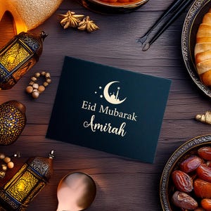 May include: A dark teal greeting card with the words "Eid Mubarak Amirah" in elegant script, adorned with a crescent moon and stars. The card is surrounded by traditional decorations, including lanterns, dates, and pastries, on a wooden surface.