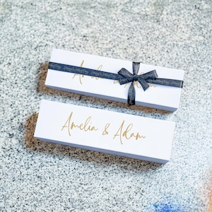 May include: Two white rectangular gift boxes. The top box has a black ribbon tied in a bow. The ribbon is printed with the words "Happy Wedding Day". The boxes have gold lettering that reads "Amelia & Adam".