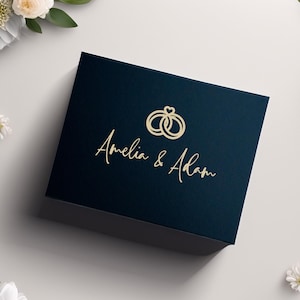 May include: A navy blue box with gold text that reads "Amelia & Adam" and a gold ring design. White flowers and greenery surround the box on a white background. The box is likely for a wedding or engagement.
