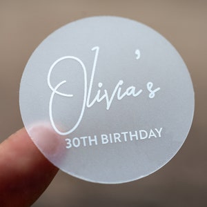 Może przedstawiać: Biała, okrągła naklejka z tekstem "Olivia's 30th Birthday" w ozdobnym, pisanym stylu.