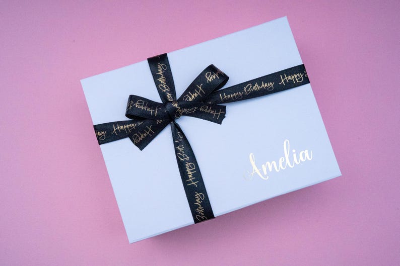 May include: A white gift box tied with a black ribbon printed with "Happy Birthday" in gold. The box is personalized with the name "Amelia" in gold script. The background is a pink surface.