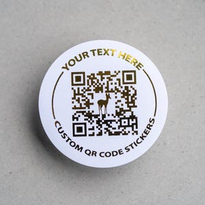 Op de afbeelding: Ronde witte sticker met gouden tekst en een QR-code. De tekst luidt "YOUR TEXT HERE" en "CUSTOM QR CODE STICKERS". Een silhouet van een hert staat in het midden van de QR-code.