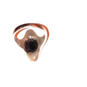 May include: A gold-toned ring with a dark, possibly black, central stone. The ring's setting has a unique, stylized shape, and the band is smooth and polished. The ring is set against a white background.