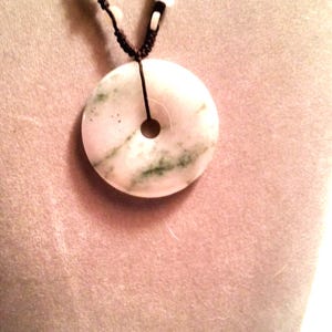 May include: A white jade pendant with green swirls and a hole in the center, hanging from a brown cord with white and orange beads.