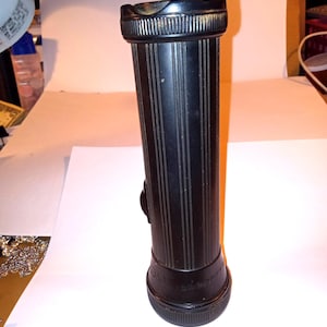 May include: A black vintage flashlight with vertical grooves and a brass handle. The flashlight has a cylindrical body and a textured base. The word "cadmium" is visible on the base. The flashlight is designed for handheld use.
