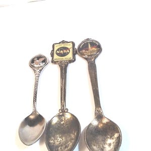 May include: Three silver souvenir spoons with different designs. The first spoon has a picture of an eagle, the second spoon has a picture of the NASA logo and the words "Space Center Houston, Texas", and the third spoon has a picture of a rocket launching.