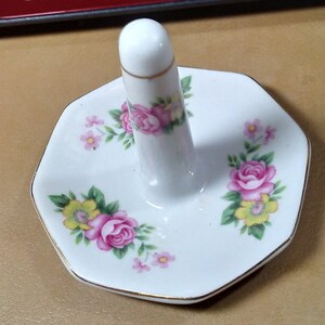 May include: White porcelain ring holder with a raised center and a gold trim. The holder is decorated with pink and yellow roses and green leaves.