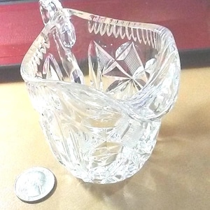 May include: A clear, cut-glass bowl with a decorative, geometric pattern. The bowl has a unique, curved shape and is likely used for serving or display. A quarter is next to the bowl for size comparison.