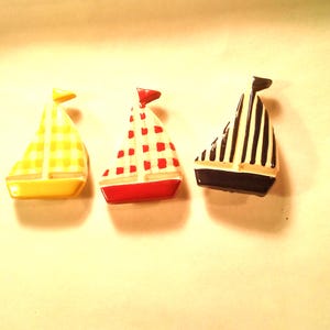 May include: Three ceramic sailboat buttons with a white background and different colored and patterned sails. The sailboats are yellow with a yellow and white checkered pattern, red with a red and white checkered pattern, and blue with black stripes.