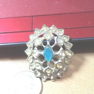 May include: A silver oval brooch with a blue stone in the center and a decorative design of smaller stones around it.