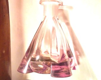 May include: A pink glass perfume bottle with a flared, pleated design. The bottle has a rounded top and a wide, geometric base. The glass has a slight iridescence, reflecting light. The bottle is likely vintage.