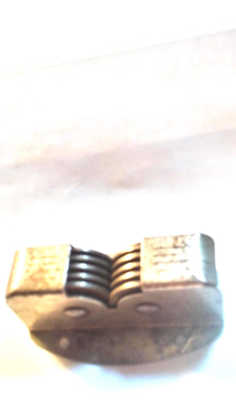 May include: A vintage metal nutcracker with a spring mechanism. The nutcracker has a rectangular body with a series of ridges and a hinged design. The metal has a tarnished appearance, suggesting age and use.