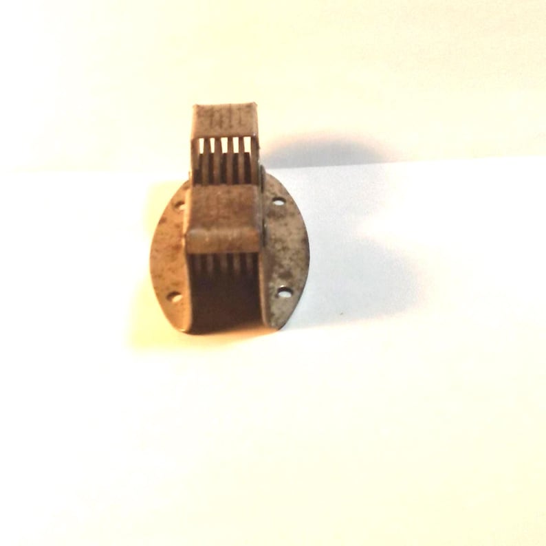 May include: A vintage, rusty metal hinge with a slotted design. The hinge is attached to an oval-shaped base with several holes for mounting. The metal has a weathered appearance, suggesting age and use. The hinge is likely for a small box or cabinet.