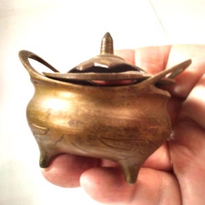 May include: A small, antique brass incense burner with a lid and three legs. The burner has two handles and a decorative design on the body. The lid has a pointed top. The overall color is a warm, aged brass.