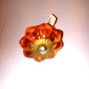 May include: An orange glass cabinet knob with a flower-like design. The knob has a gold-colored center and a clear, reflective surface. A small white tag is attached to the knob.