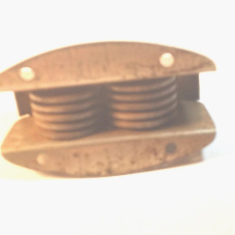 May include: A vintage, rusty metal spring assembly with two coiled springs. The assembly has a rectangular shape with rounded edges and small holes, likely for mounting. The springs are visible between the metal plates.