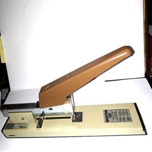 May include: A vintage Boston 135 stapler with a tan lever and base. The stapler has a silver metal mechanism and a black base. The stapler is designed for heavy-duty use.