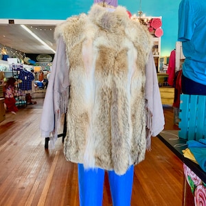 May include: A faux fur vest with a brown and white pattern. The vest has a suede-like fabric on the sides and fringe trim. The vest is displayed on a mannequin wearing blue trousers.