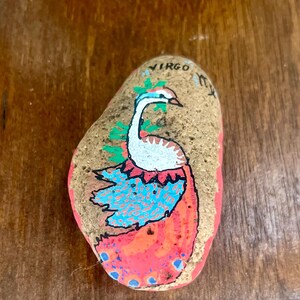 May include: Hand-painted rock featuring a peacock design with blue, orange, and white colors. The word "VIRGO" is written at the top. The rock is on a wooden surface.