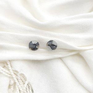 May include: A pair of small, round earrings with a white and black marbled design. The earrings are on a white fabric background.