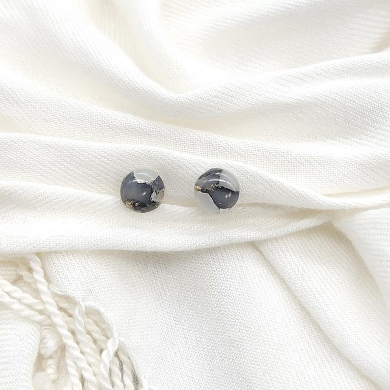 May include: A pair of small, round earrings with a white and black marbled design. The earrings are on a white fabric background.