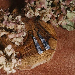 May include: A pair of silver dangle earrings with a gray and white agate stone. The earrings are on a wooden surface with dried flowers.