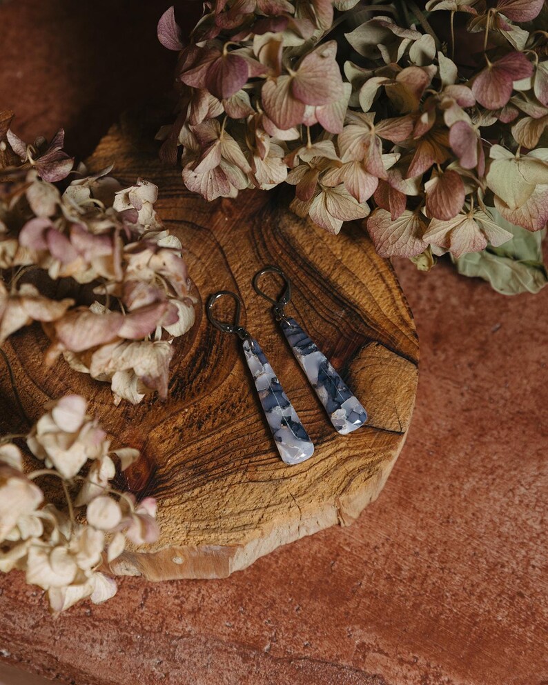 May include: A pair of silver dangle earrings with a gray and white agate stone. The earrings are on a wooden surface with dried flowers.