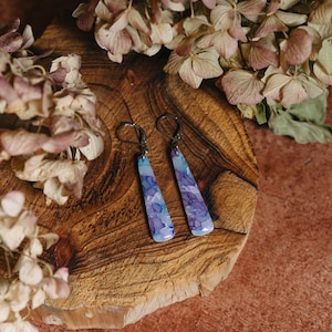 May include: A pair of dangle earrings with a purple and blue marbled design. The earrings are on a wooden surface with dried flowers.