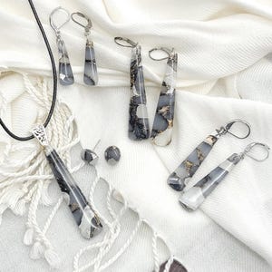 May include: A set of jewelry featuring a necklace, earrings, and a pair of studs. The jewelry is made with a white and black marbled material with gold flecks. The necklace has a long pendant and a black cord. The earrings are long and dangle, while the studs are small and round.