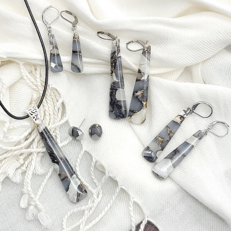May include: A set of jewelry featuring a necklace, earrings, and a pair of studs. The jewelry is made with a white and black marbled material with gold flecks. The necklace has a long pendant and a black cord. The earrings are long and dangle, while the studs are small and round.