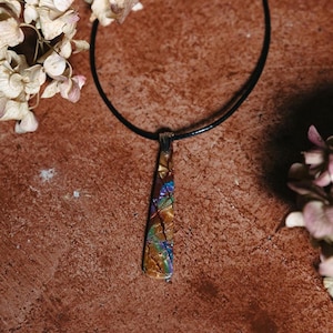 May include: A long, thin, iridescent pendant with a black cord necklace. The pendant is made of glass and has a mosaic pattern with shades of brown, gold, blue, and purple.