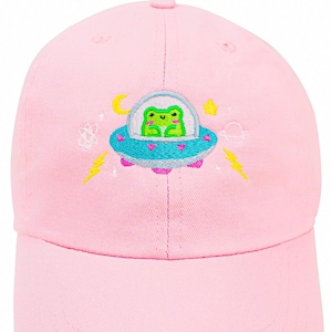 Puede incluir: Una gorra de b&eacute;isbol rosa con un dise&ntilde;o bordado de una rana verde montando en una nave espacial con corazones, estrellas y rayos.