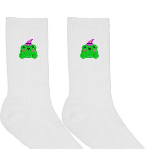 Op de afbeelding: Een paar witte sokken met een geborduurde groene kikker met een paarse tovenaarshoed op elke sok.