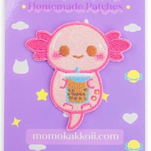 May include: Pink axolotl embroidered patch holding a cup of boba tea. The patch is on a purple background with clouds, stars, and hearts. The text "momokakkoii.com" is at the bottom of the image.