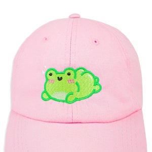 May include: A pink baseball cap with an embroidered green frog design. The frog has a happy expression and is lying on its back with its legs in the air.