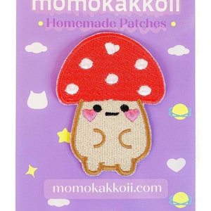 May include: A red and white mushroom with a cute face and pink cheeks. The mushroom is embroidered on a beige fabric patch. The patch is on a purple background with the text "momokakkoii.com" and the words "Homemade Patches" in white.