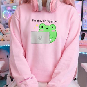 Könnte beinhalten: Hellrosa Sweatshirt mit dem Text "I'm busy on my puter" und einer grünen Frosch-Grafik, die einen Laptop hält. Auf dem Ärmel steht der Text "momokakkoii". Rosa Kopfhörer liegen auf den Schultern.