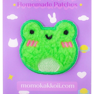 May include: Green fuzzy frog patch with pink cheeks and a white belly. The patch is on a purple background with white clouds and stars. The text "momokakkoii.com" is at the bottom of the image.